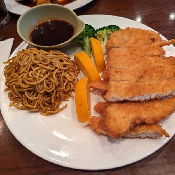 A plate of golden-brown chicken katsu, noodles, broccoli, orange slices, and a side of sauce.