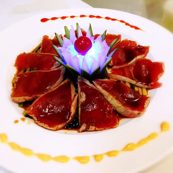 Seared tuna sashimi with red sauce, garnished with a glowing blue lotus and cherry.